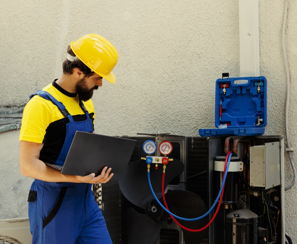 A man in blue overalls and yellow hard hat holding a laptop.